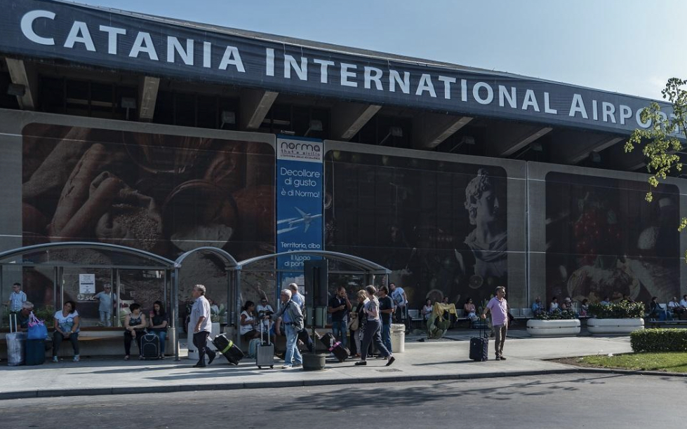 Cestujúci s kuframi pred vstupom do odletového terminálu, nad vchodom veľký nápis Catania International Airport, zamračený deň.
