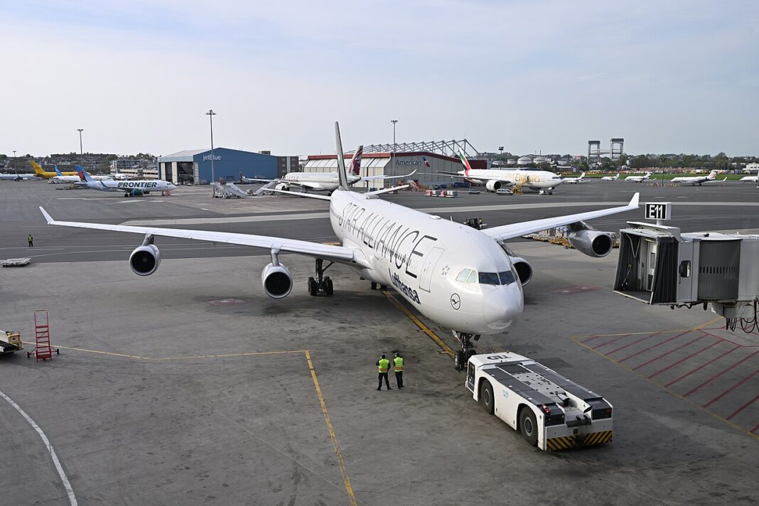 Štvormotorový Airbus A340-300 spoločnosti Lufthansa v livreji Star Alliance počas odtláčania od nástupného mosta, v popredí ťahač a dvaja pracovníci pozemnej obsluhy.