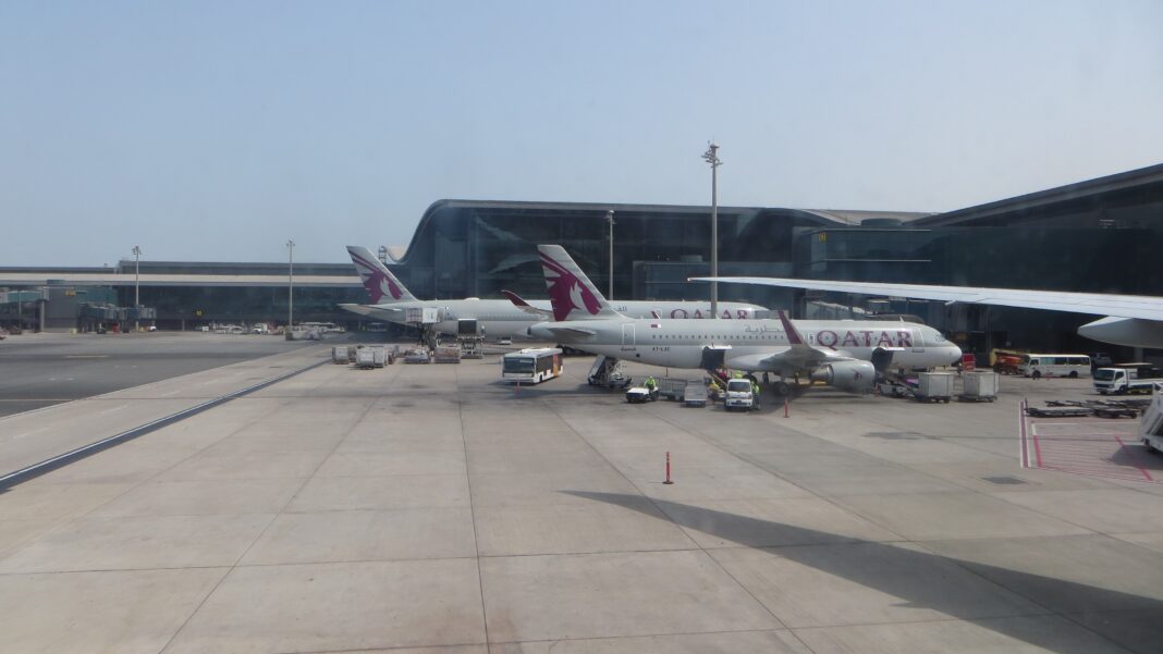 Tri lietadlá Qatar Airways stoja pri prstoch terminálu, v popredí prázdna odbavovacia plocha a vozidlá pozemnej obsluhy.