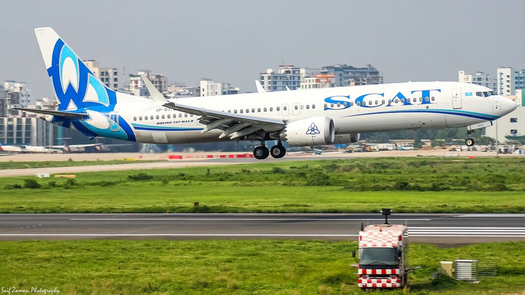 Biely úzkotrupý Boeing 737 MAX s modrým pruhom a modrým znakom na chvoste nízko nad vzletovou dráhou za denného svetla, v pozadí zelený trávnatý pás a mestská zástavba