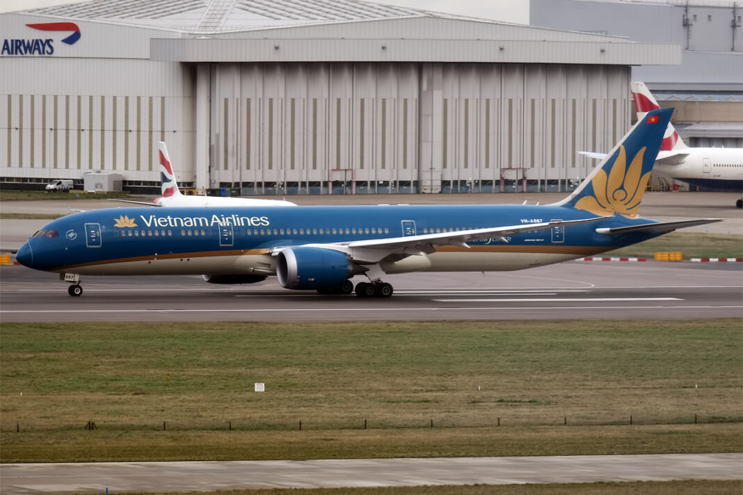 Tmavomodrý Boeing 787-9 Dreamliner spoločnosti Vietnam Airlines pri rolovaní po letiskovej ploche za denného svetla, v pozadí hangár a chvost ďalšieho lietadla