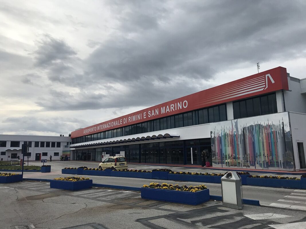 Odbavovacia budova letiska v Rimini s červeným pruhom a nápisom na fasáde pod zamračenou oblohou.